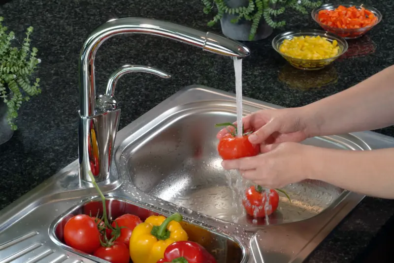 The image shows a person washing fresh tomatoes in a modern stainless steel kitchen sink equipped with a pull-out faucet. The faucet has a pull-out mousseur spray head, which extends the reach of the tap spout for convenience. Washing fruits and vegetables under running water helps to remove potential pesticides and residues. Various types of sinks and faucets are available with features like water flow limiters to save water consumption. Other vegetables, including bell peppers, are visible in the sink's smaller basin and in bowls on the counter.